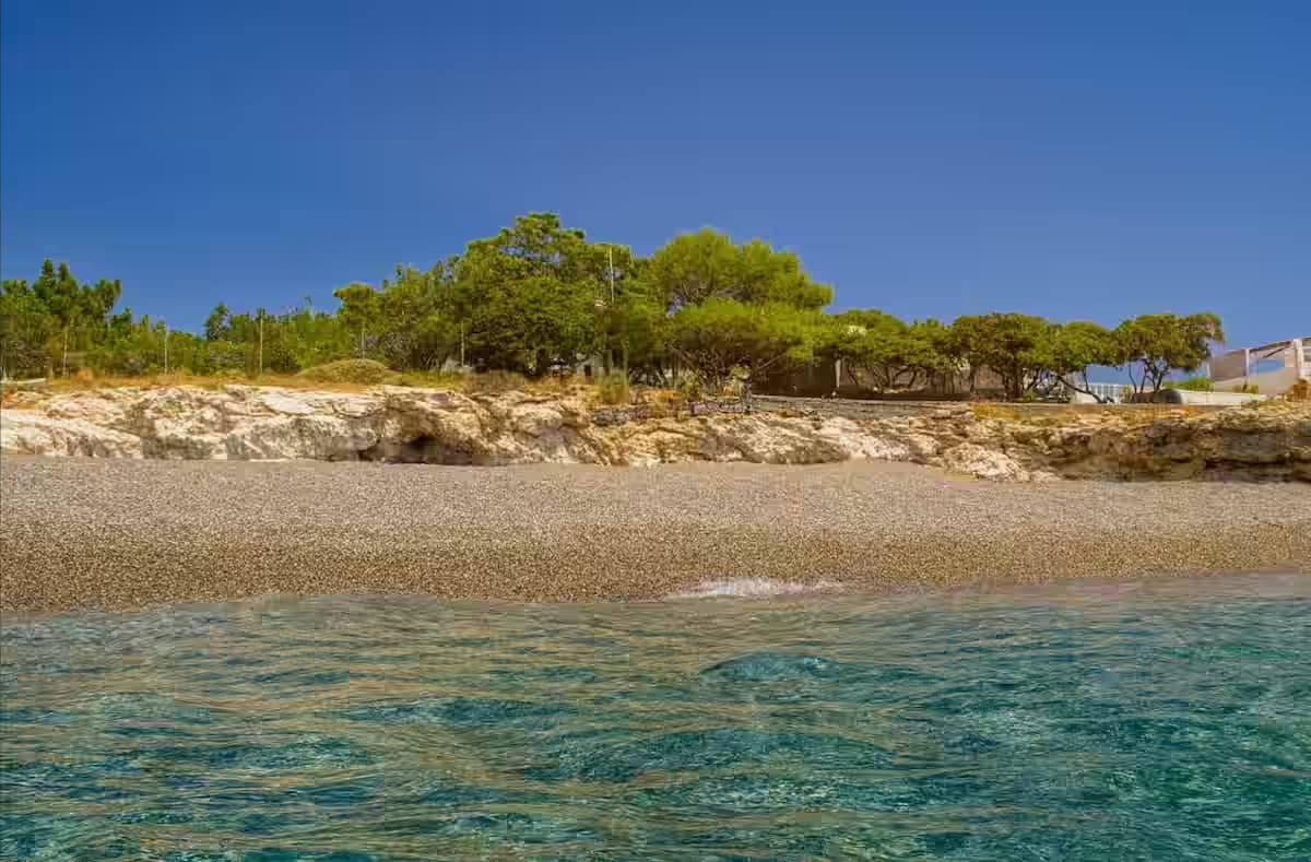 Koutsounari Long Beach, Crete, with small colorful pebbles, turquoise water, rocky embankment, and green tamarisk trees under a bright blue sky.