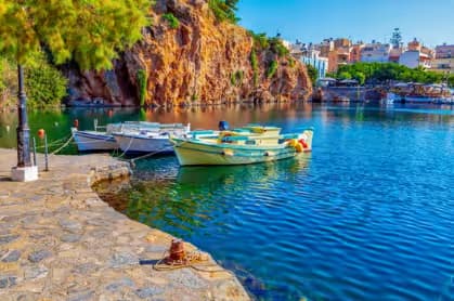 Fishing boats docked at the stone waterfront of Lake Voulismeni, Agios Nikolaos, with colorful buildings and cliffs in the background, East Crete, Greece