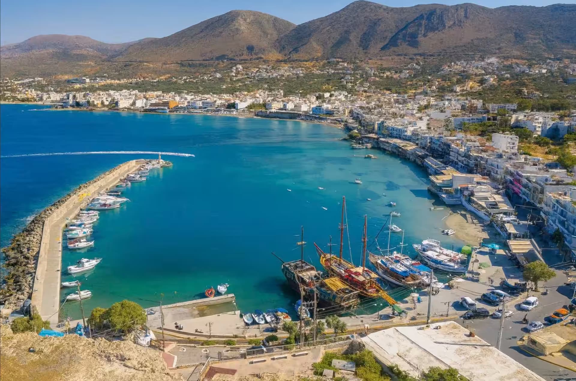 Aerial panoramic view of Hersonissos Old Port and waterfront promenade, Crete, featuring clear turquoise water and boats
